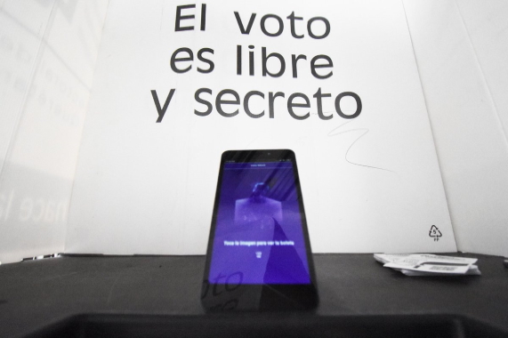 Se muestra dentro en una mampara 1 teléfono celular con la aplicación Voto Móvil, al fondo se muestra la leyenda El voto es libre y secreto