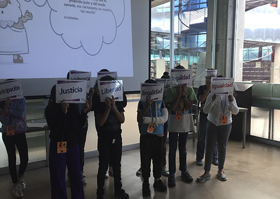 Se muestra un grupo de niños con pequeños carteles que cubren sus rostros. Los carteles contienen escrito valores como: justicia e igualdad