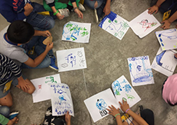 Se muestra un grupo de dibujos sobre el piso y alrededor de cada dibujo, niños que son quienes se encargan de crear la obra maestra